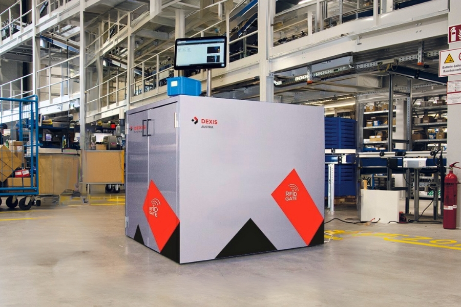Eine riesiger Behälter mit der Aufschrift „DEXIS“ und „RFID Gate“ steht in der DEXIS Austria Logistik. Das RFID-Gate hat einen Computermonitor angebracht. Der Bereich ist von Regalen, Geräten und Industriematerialien umgeben.
